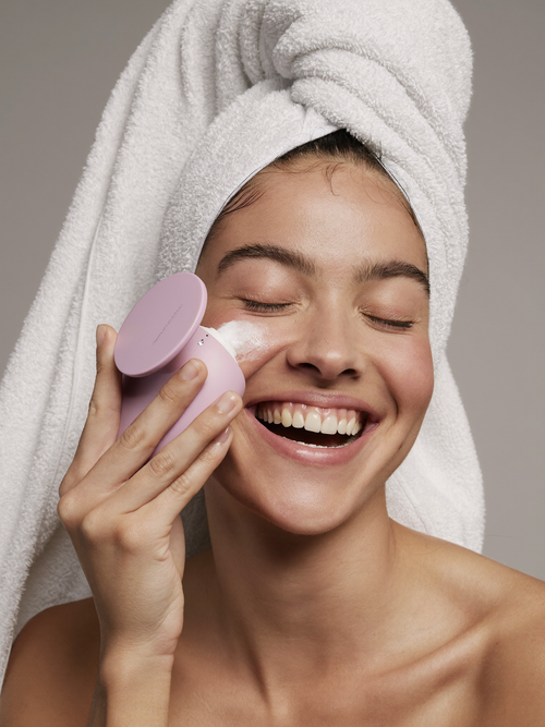 Smiling woman with a towel-wrapped head using a pink silicone facial cleansing brush